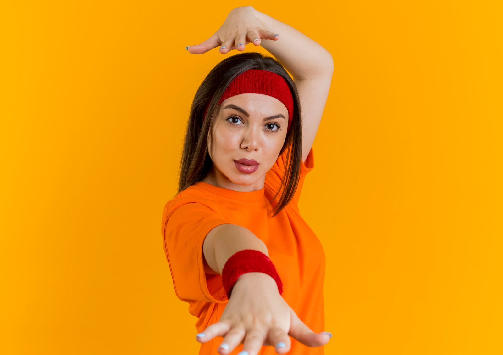 confident young sporty woman wearing headband and wristbands stretching out hand towards camera looking at camera keeping another hand above head isolated on orange background with copy space